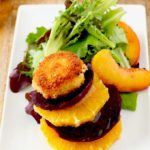 View of a stack of sliced beets and oranges topped with a crispy goat cheese coin next to salad greens and sliced peaches.