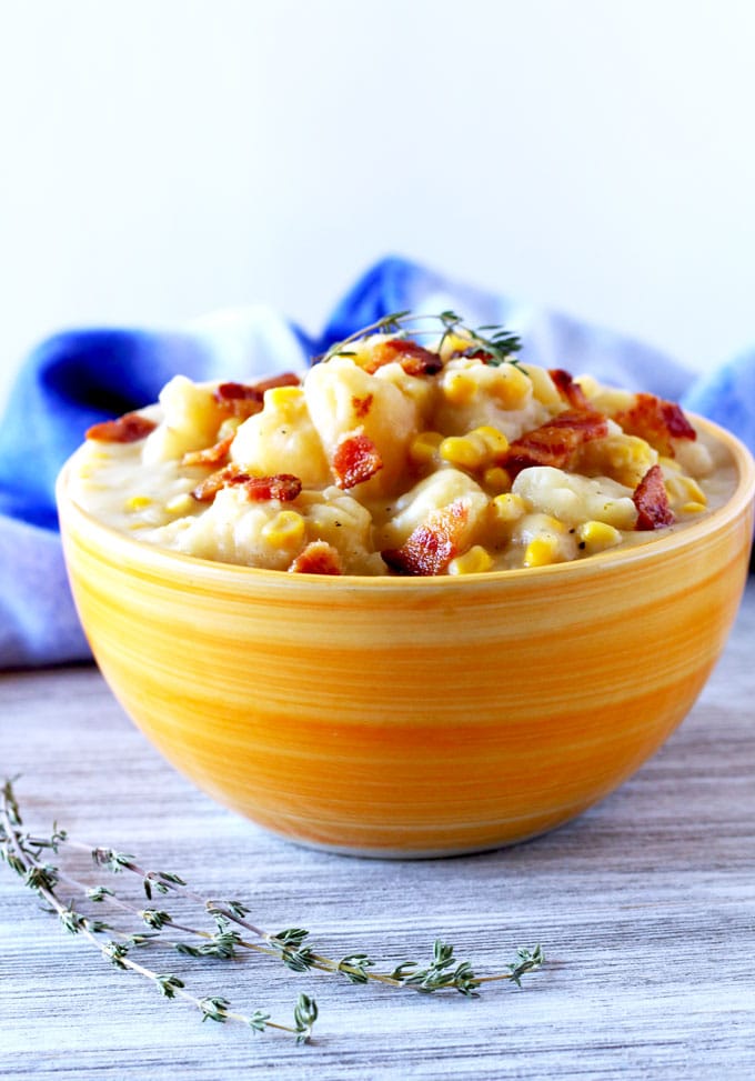 Frontal, close up view of an orange ceramic bowl filled with thick potato and corn chowder topped with crispy bacon pieces on a white wooden board. There is a blue cloth in the background.