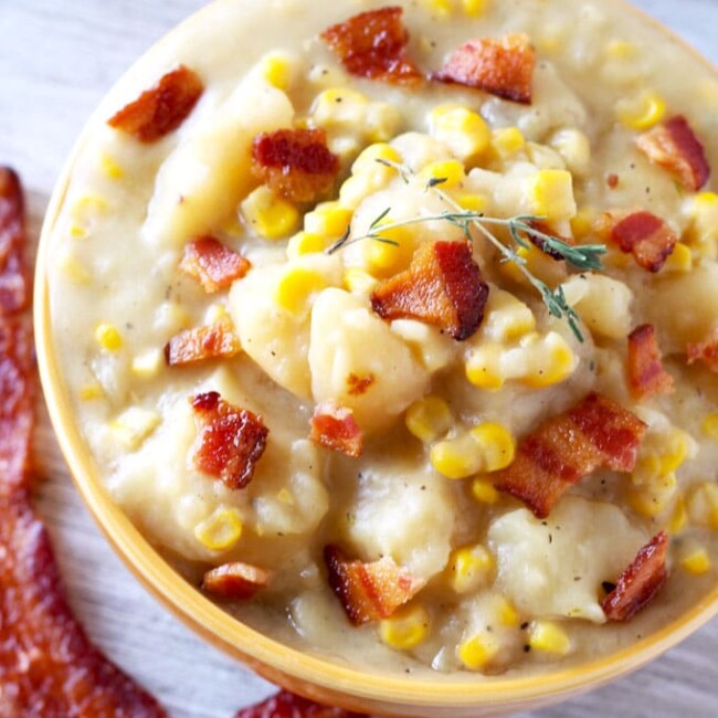 Over head, close up view of a ceramic bowl filled with thick potato and corn chowder topped with crispy bacon pieces and a couple of sprigs of thyme sitting next to a couple of slices of crispy bacon on a white wooden board.