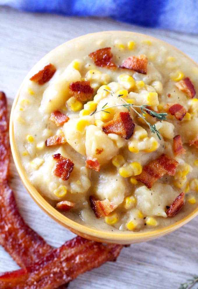 Over head, close up view of a ceramic bowl filled with thick potato and corn chowder topped with crispy bacon pieces and a couple of sprigs of thyme sitting next to a couple of slices of crispy bacon on a white wooden board.