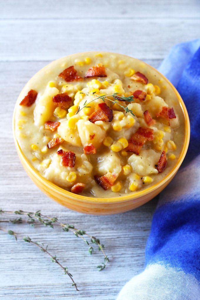 Over head, close up view of a ceramic bowl filled with thick potato and corn chowder topped with crispy bacon pieces and a couple of sprigs of thyme sitting next to a couple of thyme sprigs and a blue cloth on a white wooden board.