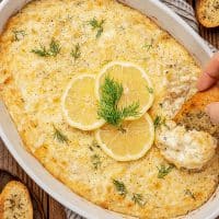 Dipping a crostini in a baking dish filled with cheesy, golden brown hot crab dip.