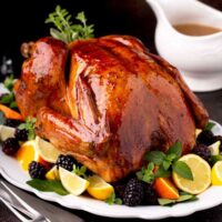 Golden brown whole roast turkey on a platter garnished with fresh herbs , citrus and fruit, next to a gravy boat.