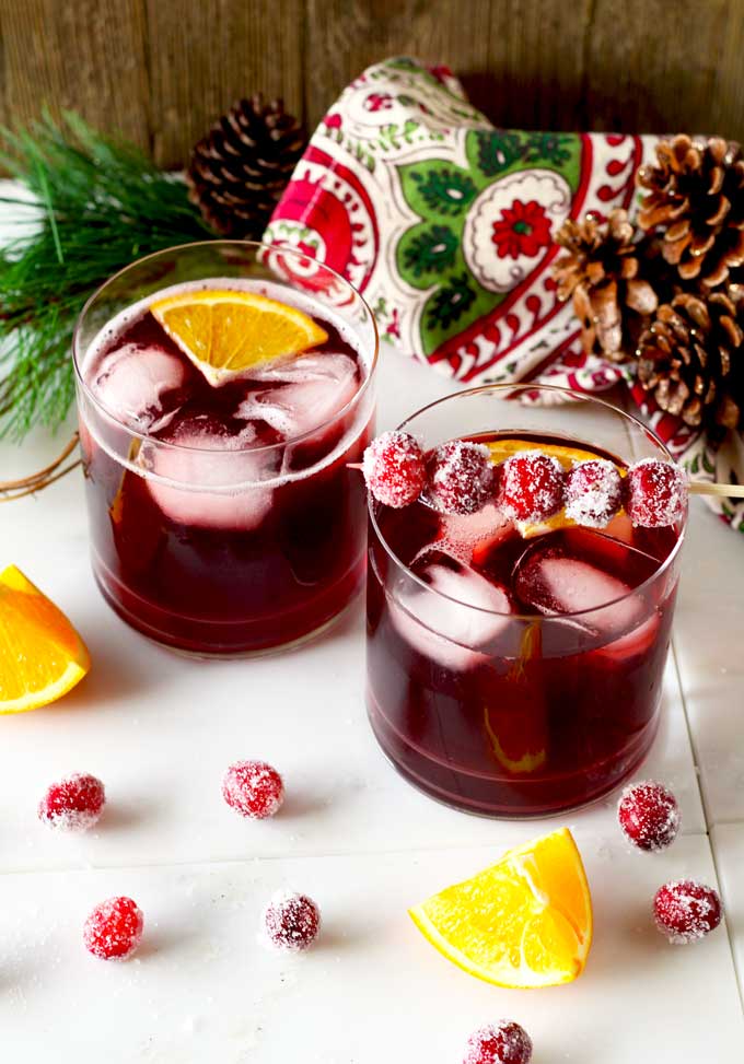 Shown here two old fashioned type glasses filled with Cranberry Maple Bourbon cocktail garnished with sugared cranberries and an orange slice.