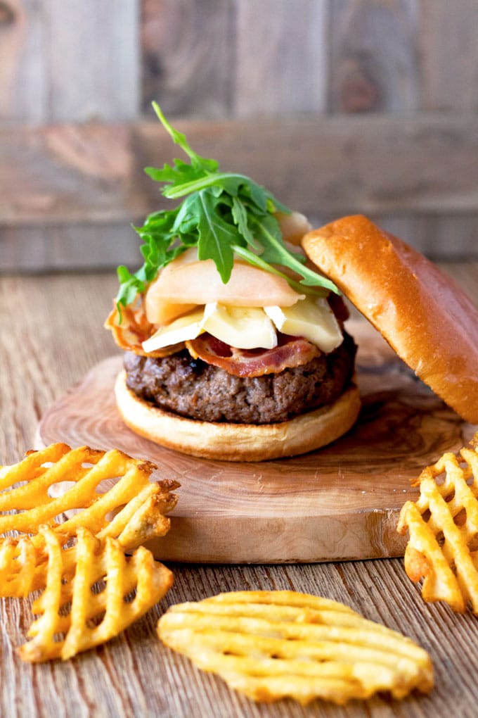 View of a burger with pancetta, brie cheese, sliced pears and arugula with the top burger bun on a side.