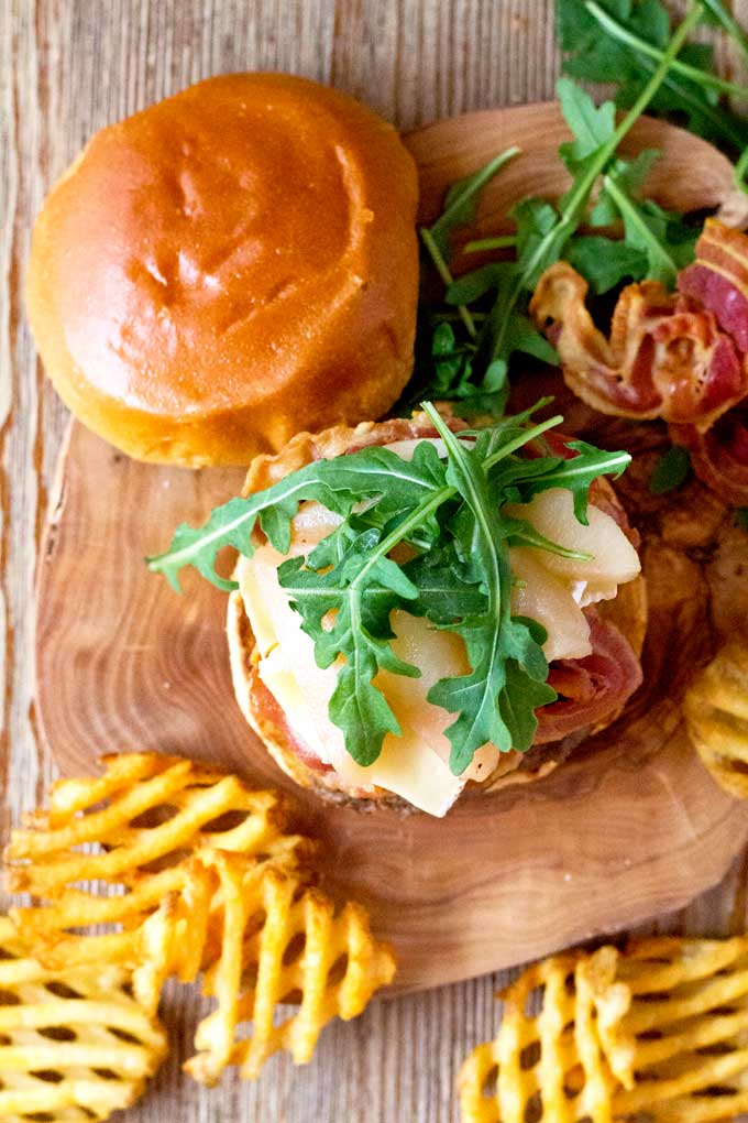 Overhead shot of a burger with pancetta, brie cheese, sliced pears and arugula in a wooden plate.