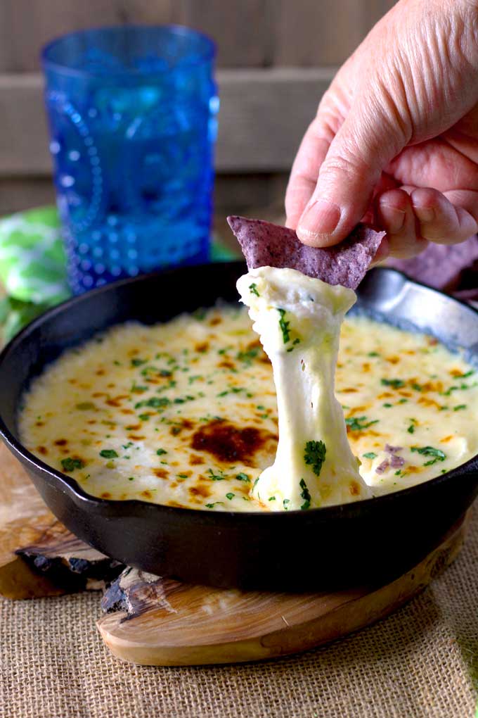 Warm and gooey Queso Fundido Dip is scooped with a blue corn tortilla chip from a cast iron skillet.