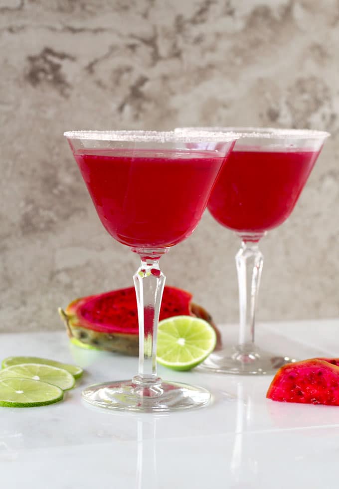 Two bright pink Margarita glasses with sugar on the rim over a white surface.