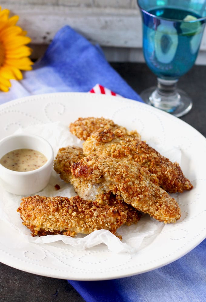 Side shot of a pile of Macadamia Nut Crusted Chicken Tenders served in a white plate with a small bowl with honey mustard