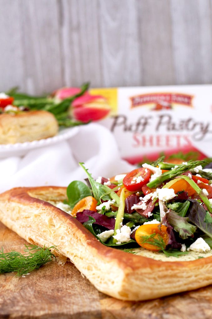 Puff pastry tart topped with greens, tomatoes, asparagus and crumbled cheese on a wooden board