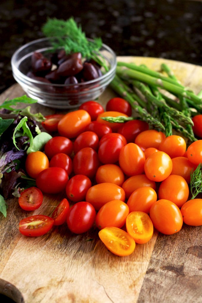 Fresh salad greens, orange and red cherry tomatoes, fresh asparagus, kalamata olives and fresh dill on a wooden surface.