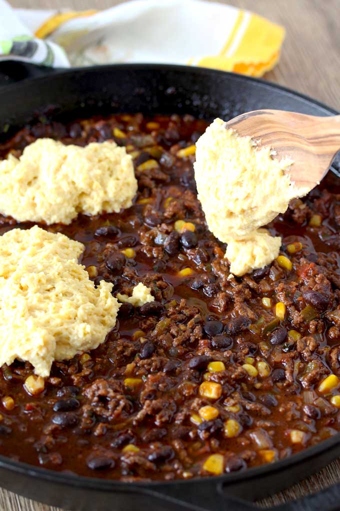 Raw Cornbread is dolloped over a mixture of ground beef chili with beans and corn, using a wooden spoon.