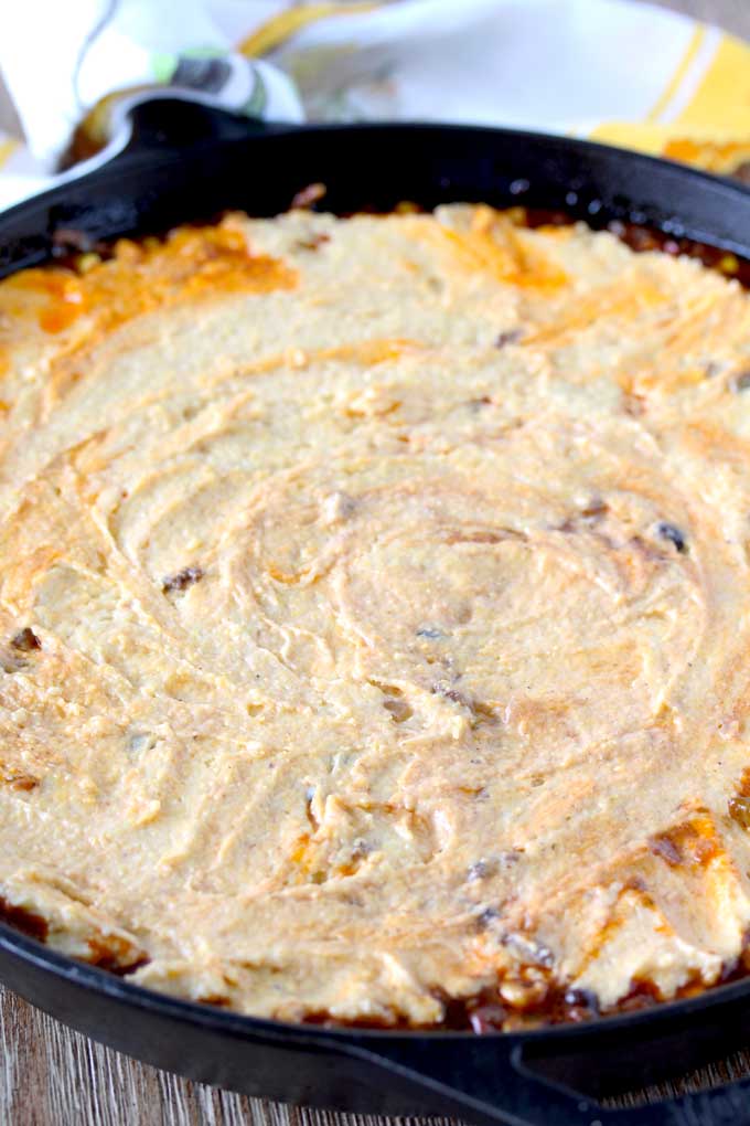 View of a skillet sitting on a wooden surface containing raw cornbread mixture spread over the top of chili. 