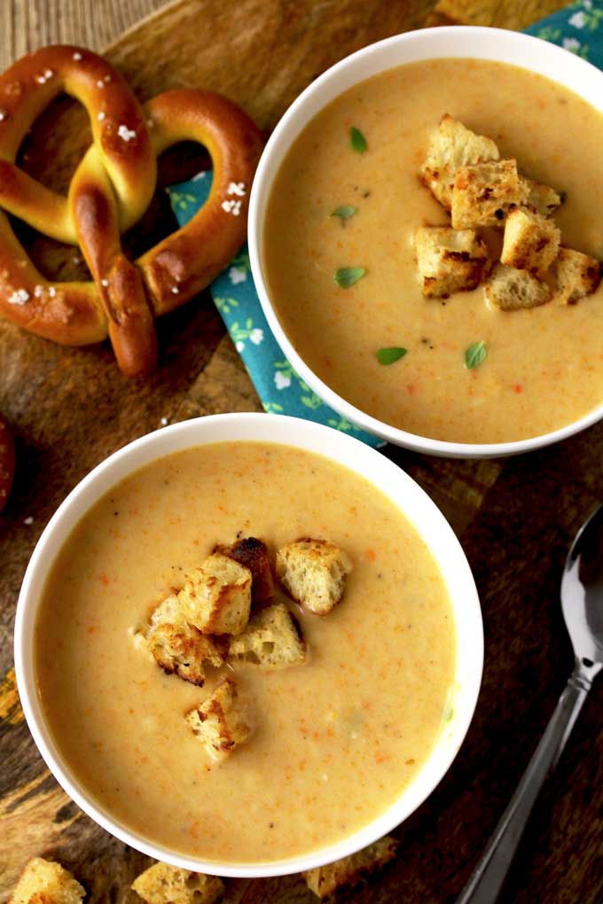 Over view of two white bowls of Cheddar Ale Soup topped with croutons and lightly garnished with thyme leaves. On the side there are two salted pretzels on a wooden board,