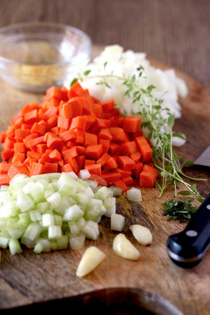 Diced celery, diced carrots, diced onions, whole garlic cloves and fresh thyme on a cutting board next to a chef's knife.