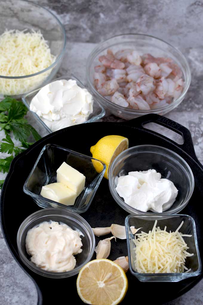 A view of different small glass bowls with ingredients. Shredded mozzarella, cream cheese, chopped shrimp and parsley on a marble surface. Next to those bowls there is a cast iron skillet. Inside there are bowls with sour cream, mayonnaise, butter, Parmesan cheese, garlic cloves and a lemon sliced in half.