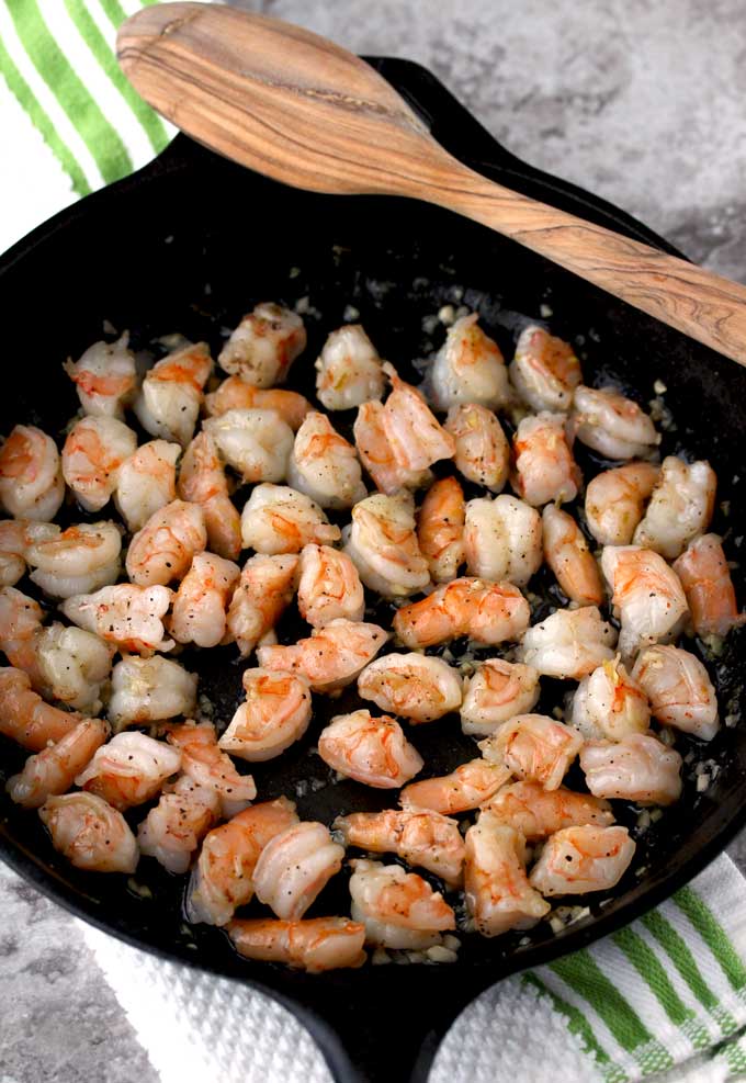 Close up view of small pieces of cooked shrimp and garlic in a cast iron skillet with a wooden spoon resting on the cast iron skillet.