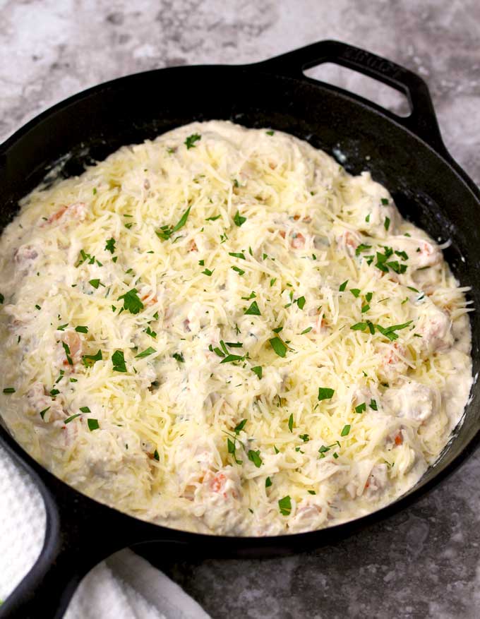 View of a cast iron skillet with the shrimp scampi dip topped with shredded mozzarella and chopped parsley ready to go into the oven,
