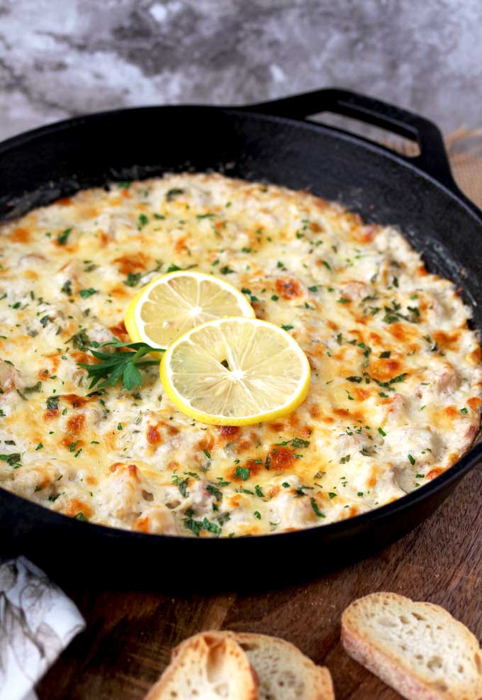 A close up view of a cast iron skillet sitting on a wooden surface filled with Shrimp Scampi Dip. The golden top is garnished with 2 thin slices of lemon and parsley. On the wooden surface you can see a few toasted croutons