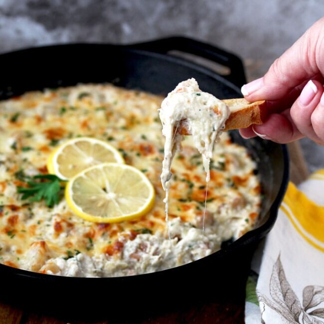 A close up view of a cast iron skillet filled with Shrimp Scampi Dip. The golden top is garnished with 2 thin slices of lemon and parsley. You can see a hand scooping the cheesy dip with a piece of toasted bread.