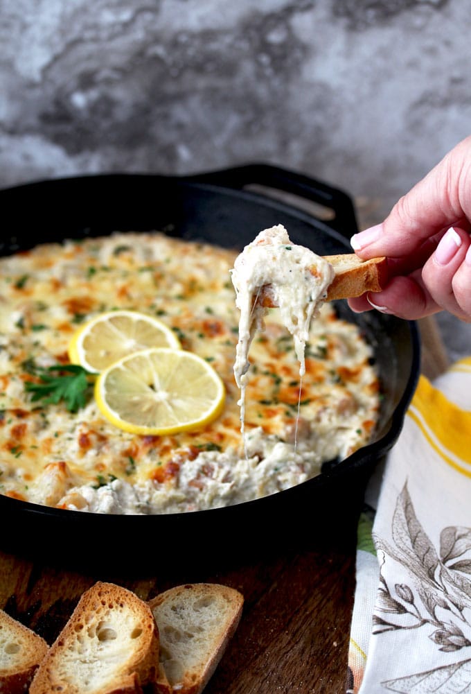 A close up view of a cast iron skillet filled with Shrimp Scampi Dip. The golden top is garnished with 2 thin slices of lemon and parsley. You can see a hand scooping the cheesy dip with a piece of toasted bread.