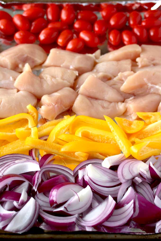 View of a sheet pan with rows of cherry tomatoes, chicken raw chicken pieces, sliced yellow bell peppers and sliced red onions.