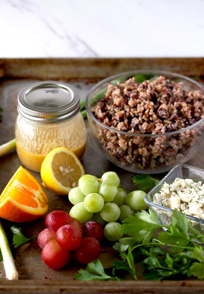 Wild Rice Salad Ingredients on a metal surface. Citrus Miso Vinaigrette in a small jar, cooked wild rice in a bowl, green and red grapes, orange, lemon, blue cheese, green scallions and parsley.