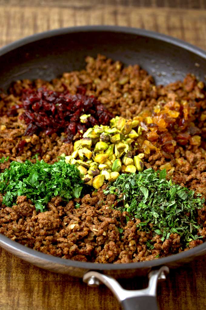 View of a skillet filled with cooked ground beef. On top, chopped, cranberries, chopped golden raisins. chopped pistachios, chopped parsley and chopped mint.
