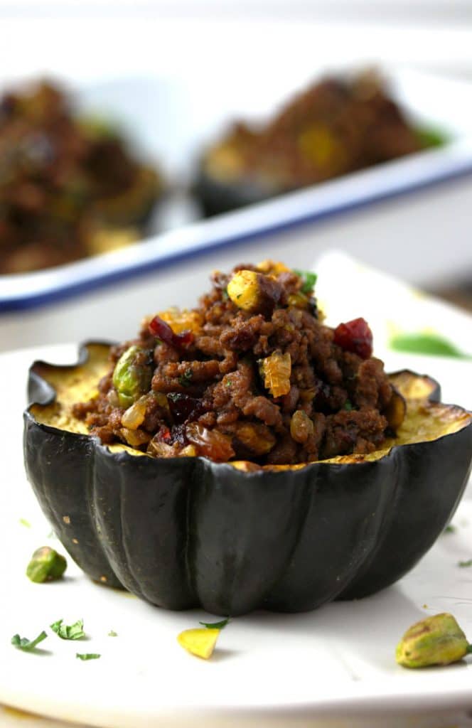 Side view of a stuffed acorn squash with ground beef, pistachios, dried cranberries, golden raisins, chopped parsley and mint on a white plate.