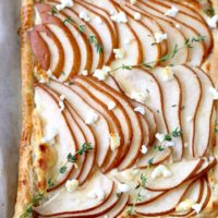 Top view of a golden brown puff pastry tart topped with thinly sliced pears garnished with goat cheese and fresh thyme on a parchment paper lined baking sheet.
