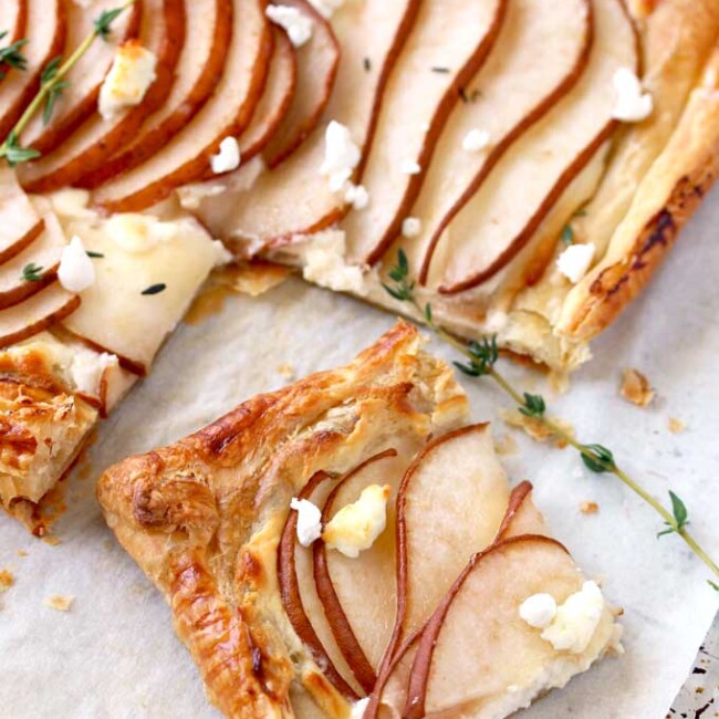 View of a golden brown puff pastry tart sliced The tart is topped with thinly sliced pears garnished with goat cheese and fresh thyme on a parchment paper lined baking sheet.
