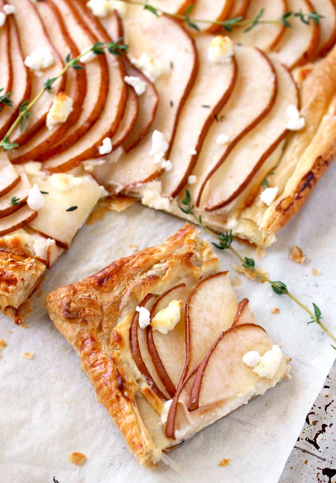 View of a golden brown puff pastry tart sliced The tart is topped with thinly sliced pears garnished with goat cheese and fresh thyme on a parchment paper lined baking sheet.