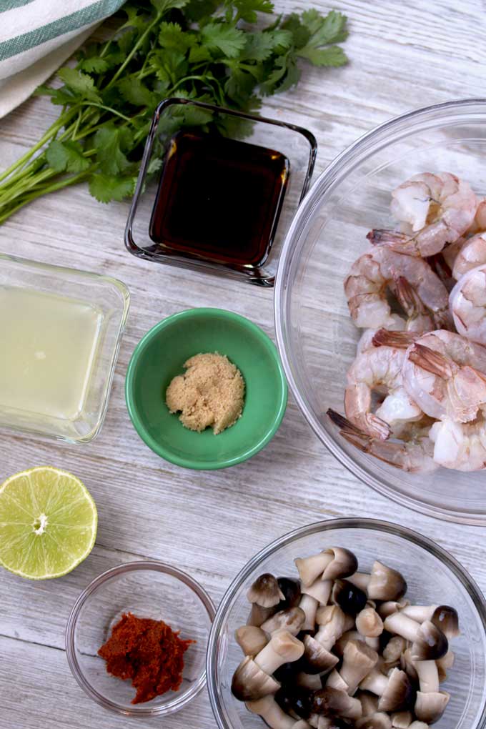 View of ingredients for Thai Tom Yum Soup on a white wooden board. Fresh raw shrimp with the tails on. Lime juice, fish sauce, brown sugar, Thai red chili paste, straw mushrooms and fresh cilantro.