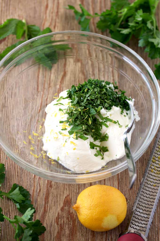 View of a glass bowl with the ingredients for making the herbed yodurt dip. Greek yougurt, chopped parsley, chopped mint and lemon zest.
