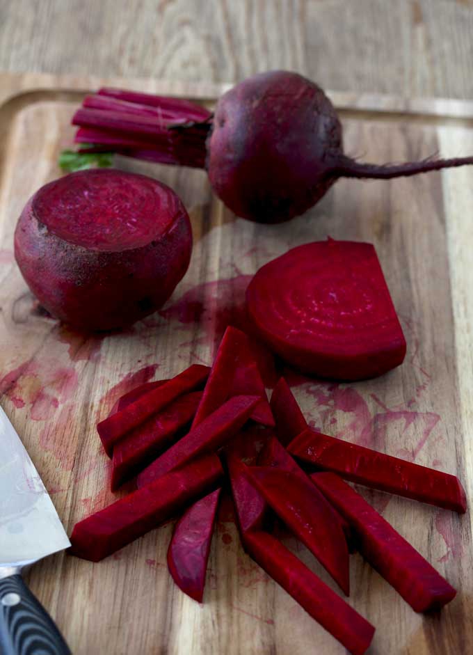 View of the process of cutting beets in the manner of French fries. A whole beet, a beet with the ends cut of and a beet that has been sliced and cut into thin strips on top of a cutting board.