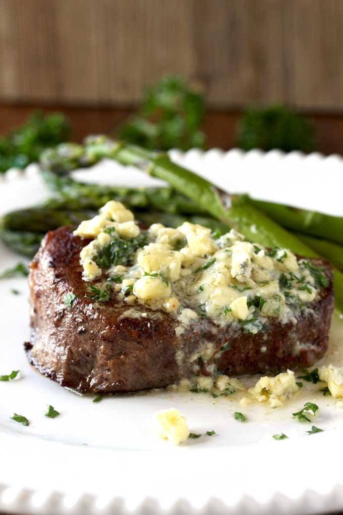 Closed up view of a pan seared filet mignon topped with blue cheese butter. The butter is melting and you can see some blue cheese crumbles and chopped parsley sitting on a white plate. Next to the filet mignon, sauteed asparagus.