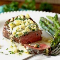 Close up view of a perfectly seared filet mignon topped with melty blue cheese butter. It has been sliced thinly and it is cooked to medium rare temperature. Also on the plate, sauteed asparagus.