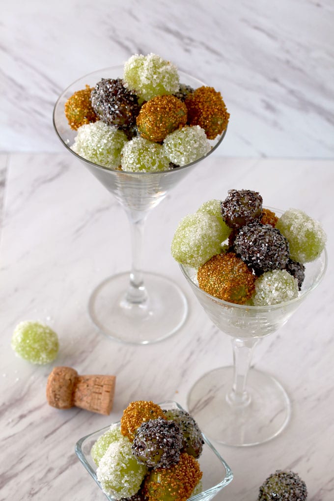 Top view of three crystal containers filled with sugared grapes. Also a few grapes and a Prosecco cork on a white marble surface.