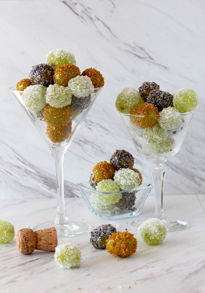 View of three crystal glasses filled with Prosecco Sugared Grapes covered in silver, gold and sparkling white sugars on a white marble surface.