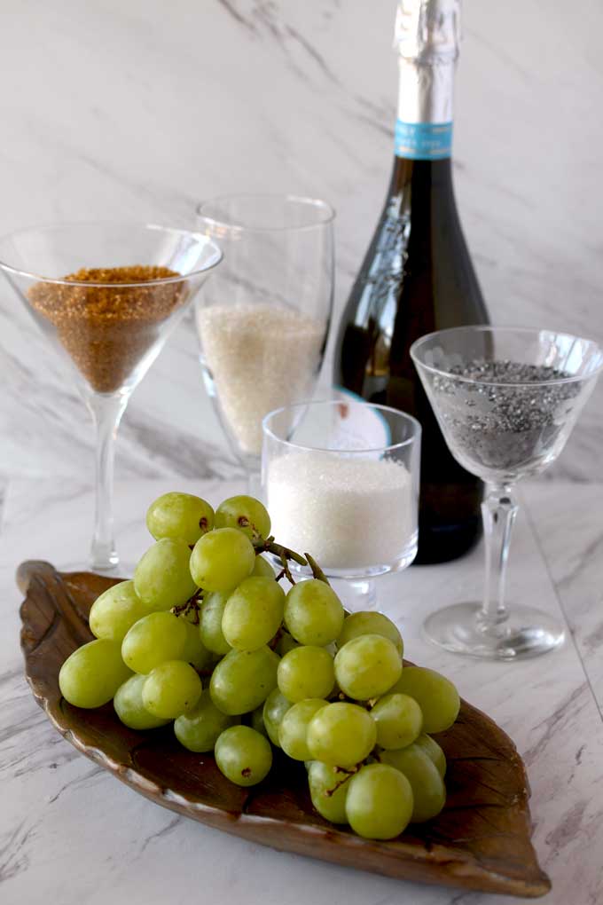 Ingredients for making Prosecco Sugared Grapes sitting on a white marble counter. A bunch of greenn grapes, sanding sugars and a bottle of Prosecco.