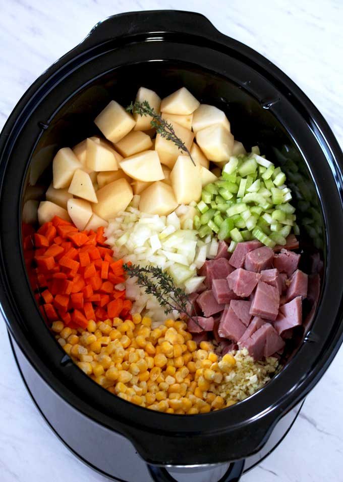 Pictured here is a slow cooker filled with cubed potatoes, chopped onions, celery, carrots, corn, minced garlic and thyme sprigs.