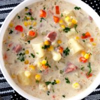 Top view of a white bowl filled with creamy ham and potato soup with potatoes, corn and carrots and garnished with chopped parsley sitting on a striped black and white table mat.