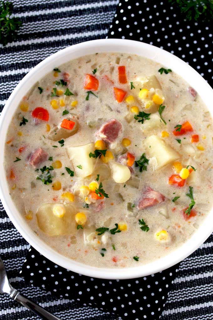 Top view of a white bowl filled with creamy ham and potato soup with potatoes, corn and carrots and garnished with chopped parsley sitting on a striped black and white table mat.