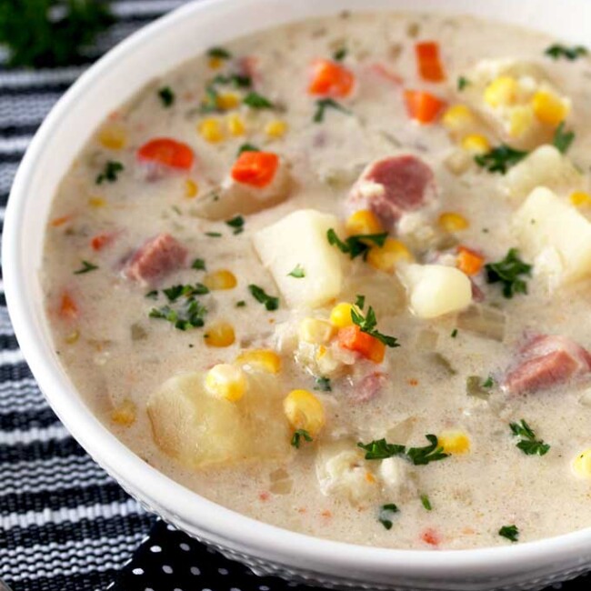 Pictures a big bowl of creamy slow cooker ham and potato soup with diced ham, potatoes, carrots and corn on a black and white table mat.