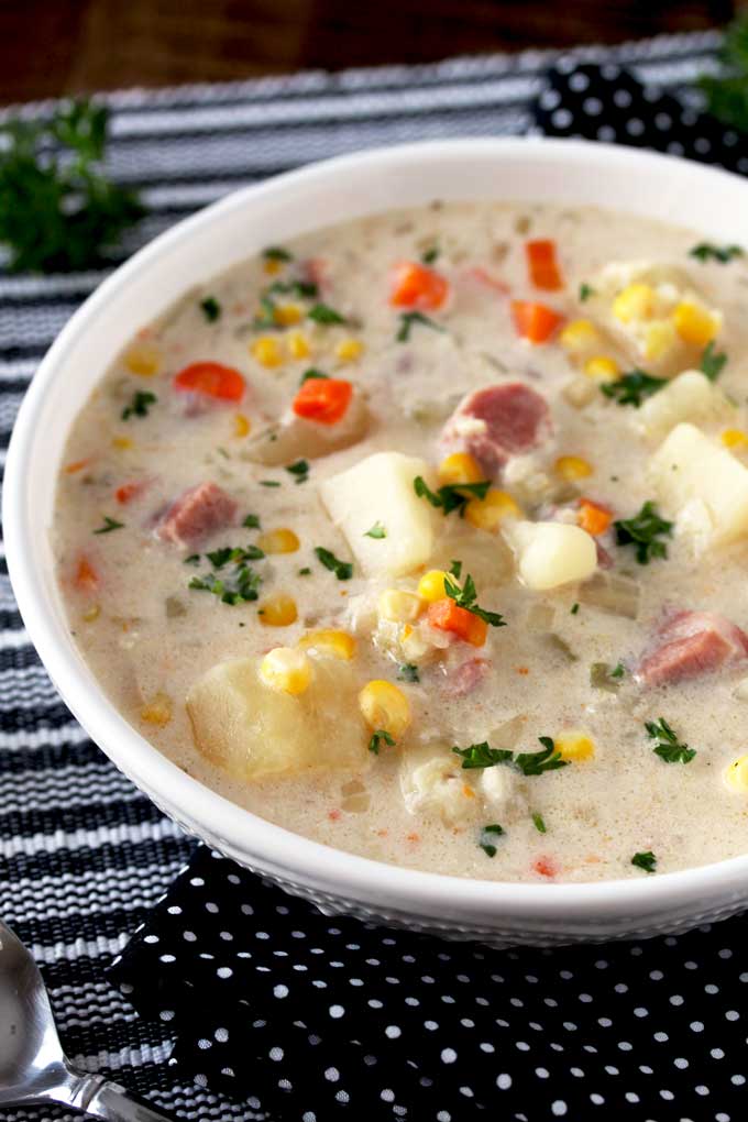 Pictures a big bowl of creamy slow cooker ham and potato soup with diced ham, potatoes, carrots and corn on a black and white table mat.