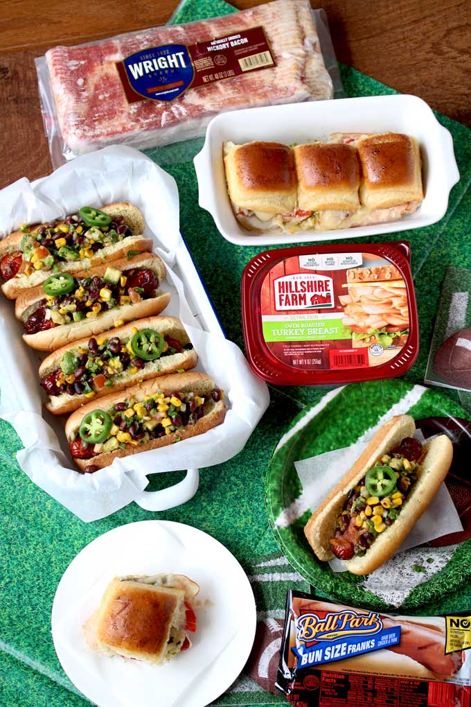 Game Day Foods set up over a green tablecloth. Southwest Bacon Wrapped Hot Dogs on a tray garnished with cilantro and jalapeno slices and pesto turkey sliders. Also a package of sliced turkey, hot dogs and bacon.