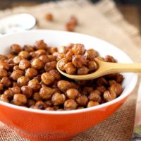 Pictured here is a small orange bowl filled with crispy oven roasted chickpeas. A small wooden spoon si scooping out some chickpeas from the bowl.