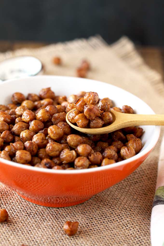 Pictured here is a small orange bowl filled with crispy oven roasted chickpeas. A small wooden spoon si scooping out some chickpeas from the bowl.
