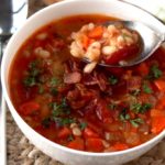 A spoon is scooping Bean and Bacon Soup from a white bowl.