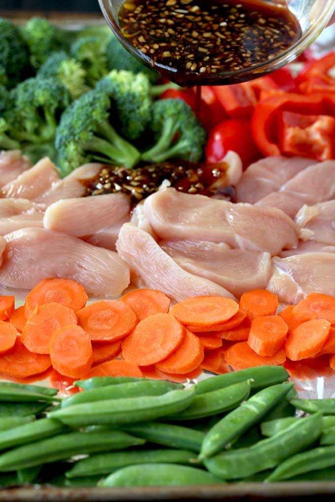 Pictured here a small bowl with stir fry sauce is been poured over a sheet pan with ingredients to make this easy Asian Chicken and Vegetables Stir Fry. Raw chicken, sliced carrots, red bell peppers, broccoli florets and sugar snap peas.